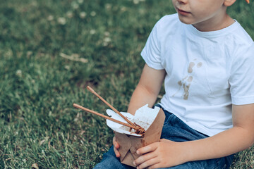 The boy holding takeaway food sitting on the grass. Dirty sauce stains on a white t-shirt. outdoors