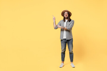 Fototapeta premium Full body happy young man he 20s in striped grey shirt white t-shirt hat pointing index finger aside indicate on workspace area copy space mock up isolated on plain yellow background studio portrait