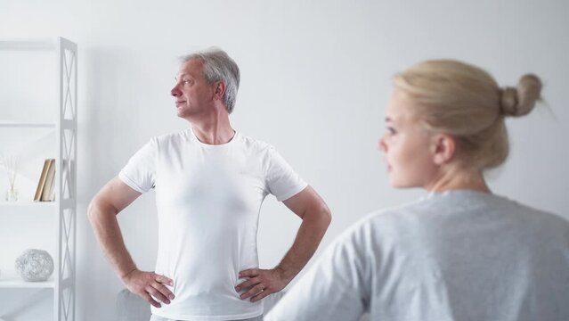 Personal training. Fitness class. Morning exercise. Inspired middle-aged man stretching neck muscles with female coach in light home studio interior.