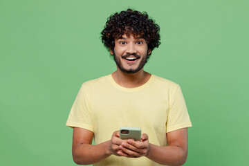Young excited happy Indian man 20s in basic yellow t-shirt hold in hand use mobile cell phone...