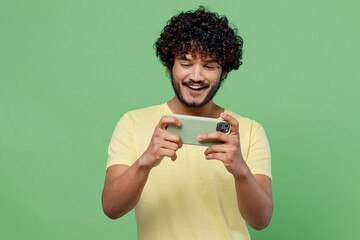 Full body young gambling happy Indian man 20s in yellow t-shirt using play racing app on mobile cell phone hold gadget smartphone for pc video games isolated on plain pastel light green background © ViDi Studio
