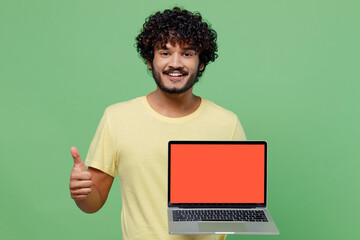 Young cool fun happy Indian man 20s in basic yellow t-shirt hold use work on laptop pc computer with blank screen workspace area show thumb up isolated on plain pastel light green background studio