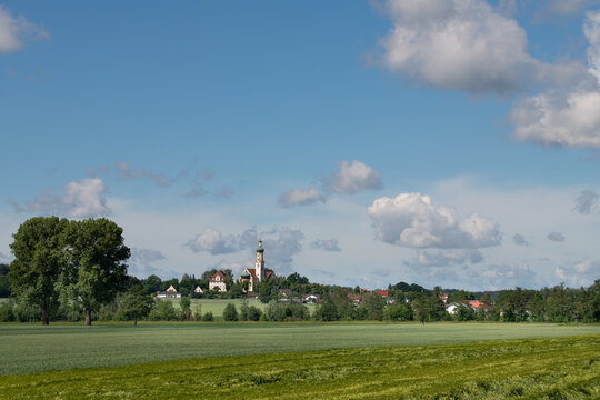 Landscape shot in Bavaria. On the horizon the pilgrimage church of Biberbach. In the foreground trees and a green field of wheat. In the blue sky are white clouds.