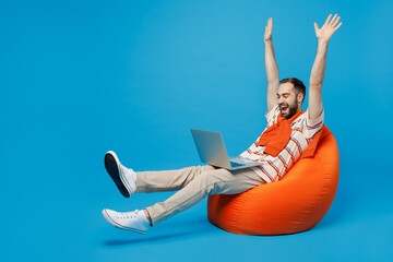 Full body young excited fun cool man 20s in orange striped t-shirt sit in bag chair hold use work on laptop pc computer with overstretched hands legs isolated on plain blue background studio portrait - obrazy, fototapety, plakaty