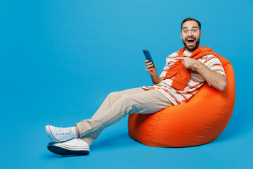 Full body young smiling fun happy man 20s in orange striped t-shirt sit in bag chair hold point...