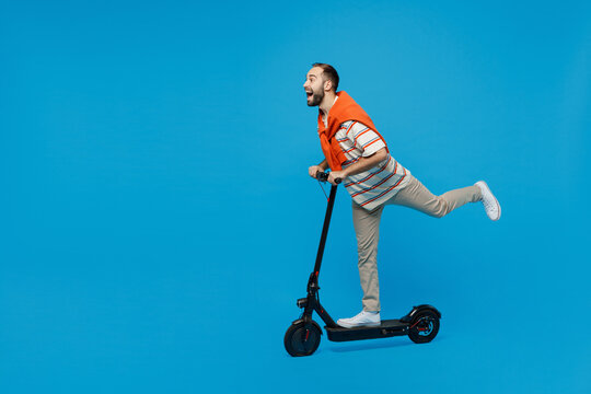 Full Body Side View Young Happy Caucasian Man 20s Wearing Orange Striped T-shirt Riding Electric Scooter Raise Up Leg Push Isolated On Plain Blue Background Studio Portrait. People Lifestyle Concept.