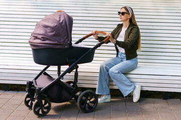 A young mother is sitting on a bench in the park next to a child who is lying in a stroller