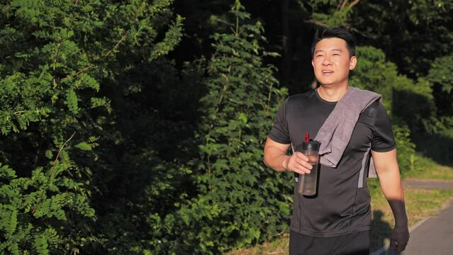 Happy asian man with a towel on his shoulder walking in summer park drinking water after running outdoor. Finishing workout, catching breath enjoys fresh air. Green trees on background.