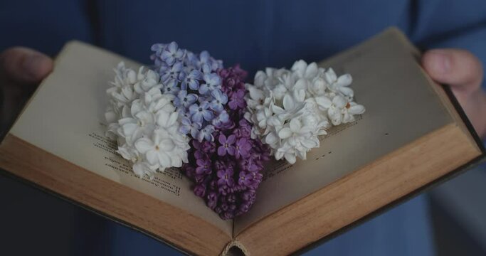 Close-up of unrecognizable women's hands opening an old book of poetry. Vivid lilac flowers bloom on its pages. The concept of romantic poetry, tenderness and dreaminess.