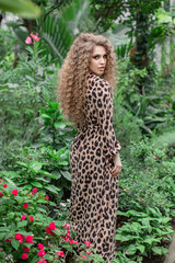A sexy girl model with a slender body in leopard print, a dress with a pattern, afro-curls and professional makeup stands in the leaves of tropical trees, plants. Summer atmosphere