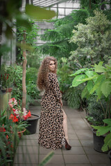 A sexy girl model with a slender body in leopard print, a dress with a pattern, afro-curls and professional makeup stands in the leaves of tropical trees, plants. Summer atmosphere
