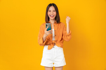 Cheerful young Asian woman holding smartphone with fists clenched celebrating victory expressing...