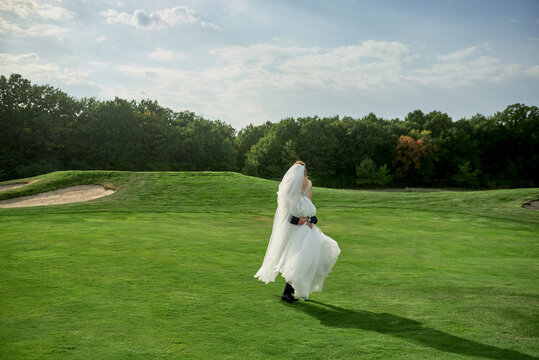 Full length body portrait of young bride and groom dancing on golf course, back view. Happy wedding couple on green grass, copy space