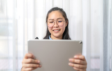 Smiling Asian Woman holds and watches gray tablet on her hand in front of curtain in private room in Work from Home period.