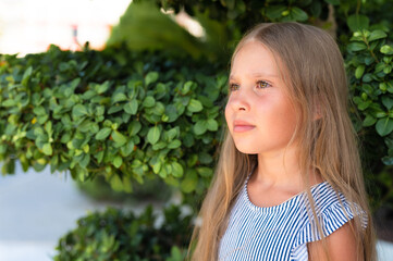 portrait face of candid happy little kid girl of eight years old with long blond hair and green eyes on background of green plants during a summer vacation travel. gen z mental health concept