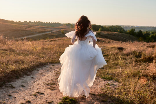 Full length body portrait of beautiful bride in white wedding dress running in the field on sunset, back view. Wedding concept