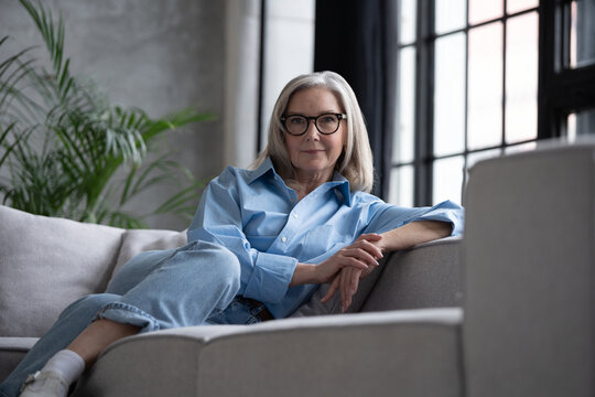 Portrait of charming senior woman with grey hair, smiling mature female sits on the couch and looks at the camera