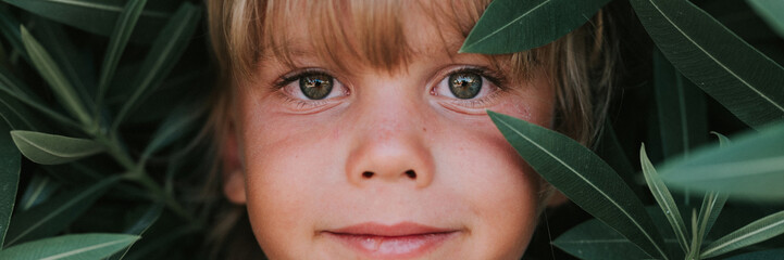 Obraz premium portrait face of candid happy little kid boy of five years old with problem allergic skin and long blond hair and green eyes on background of green plants during a summer vacation travel. banner