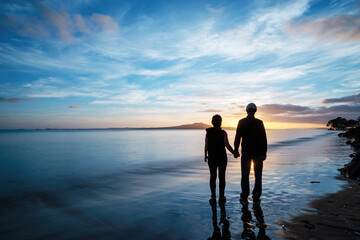 Silhouette image of a couple standing on Milford beach, watching sun rising above Rangitoto Island, Auckland. - obrazy, fototapety, plakaty