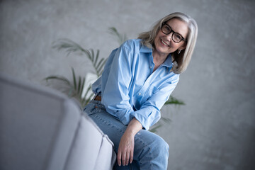 Fototapeta na wymiar Portrait of charming senior woman with grey hair, smiling mature female sits on the couch and looks at the camera