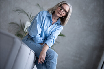 Fototapeta na wymiar Portrait of charming senior woman with grey hair, smiling mature female sits on the couch and looks at the camera
