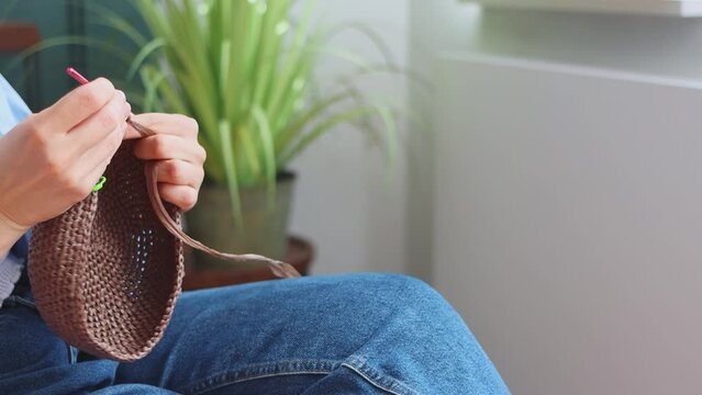 Woman knitting decorative brown basket using pink hook and raffia yarn sitting at home crocheting indoors. Creation of handmade accessories from natural materials, hobby. High quality 4k footage.