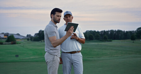 Two golfers looking tablet watching tournament game video at golf course sunset.