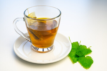 Nettle herbal tea in a transparent cup, with a sachet immersed in water, and a white saucer with nettle leaves next to it. Healing infusion of nettle. 
