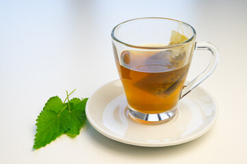 Nettle herbal tea in a transparent cup, with a sachet immersed in water, and a white saucer with nettle leaves next to it. Healing infusion of nettle. 
