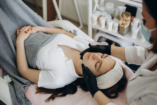 Cropped image of female beautician dripping moisturizing oil with dropper on face of girl in beauty salon. Blurred young asian woman client with closed eyes wearing medical cap. Body and skin care