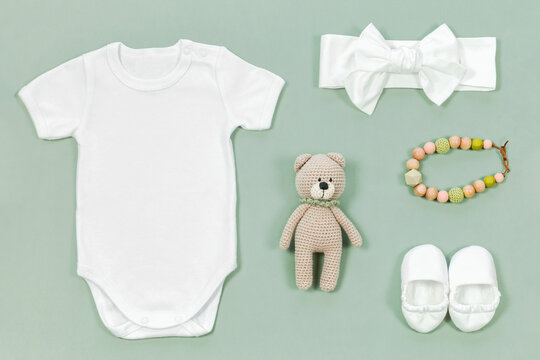 White baby bodysuit mockup on mint green pastel background with kids accessories. Teether beads, headband, booties and hand knitted teddy bear toy lie next to a romper for a mockup or design template.