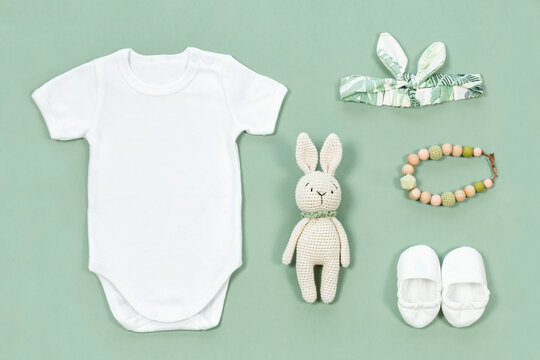White bodysuit for baby mock-up. Mockup of infant clothes on a mint green background with baby toys and accessories. Crocketed toy of rabbit, cute head band, booties and teather beads next to a romper