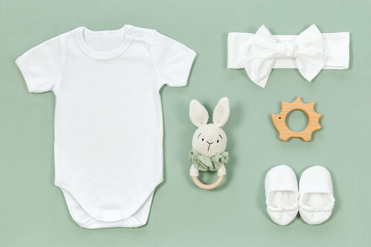 Cotton baby bodysuit for mockup on a mint green background. Infant accessories and wooden toys next to white romper for kids clothes mock-up. Cute baby headband and booties in eco style top view.