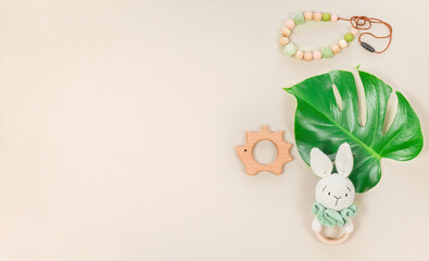 Baby accessories and toys banner with copy space in eco style with tropical monstera leaf. Lovely wooden teethers, a handmade knitted bunny, baby beads and a headband on a pastel brown background. © nieriss
