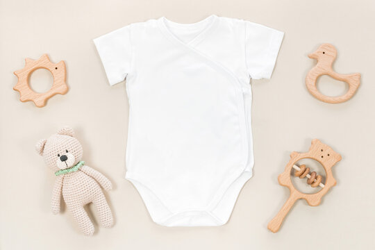 White wrap bodysuit for baby mocap. Mockup of children's clothes on a pastel brown background with kid's toys and accessories. Wooden rattles and teethers next to a cotton romper top view.