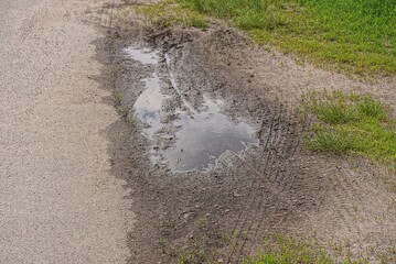 one big puddle of dirty water on the gray earth of a rural road near green grass