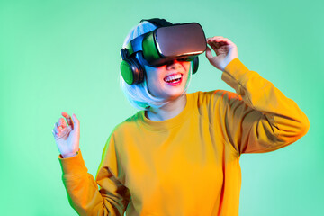 Metaverse concept, young asian woman in yellow sweatshirt wearing vr glasses playing and watching on the neon green background.