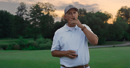 Rich golf player stand relaxed on course. Old man smoking cigar holding drink.