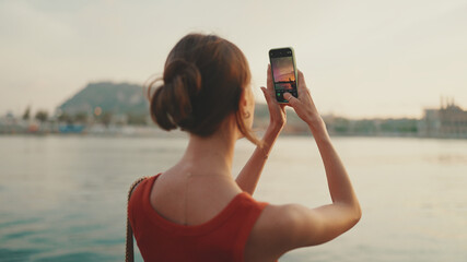 Close-up, girl stands on the embankment and takes pictures of sunrise on a mobile phone. Closeup of young woman shoots the sea on smartphone in the morning time. Back view