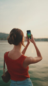 Close-up, girl stands on the embankment and takes pictures of sunrise on a mobile phone. Closeup of young woman shoots the sea on smartphone in the morning time. Back view
