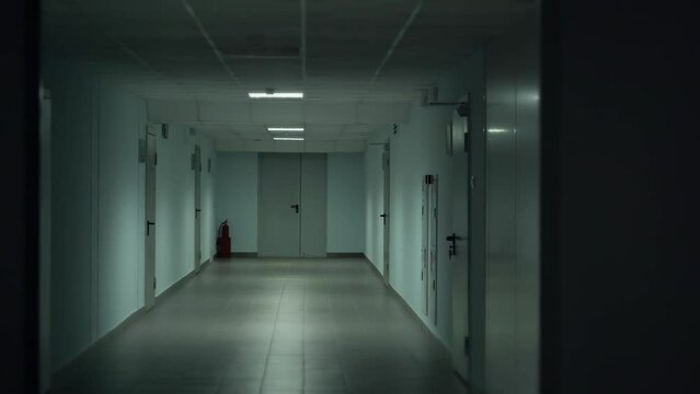Tracking inside a long dark gloomy corridor, tunnel past the small windows to the old ajar doors with glass inserts. Concept of horror.