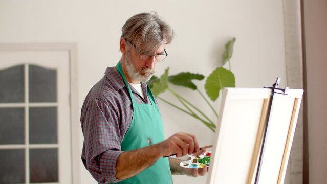 Elderly male artist in an apron thoughtfully and intently paints on canvas in living room, front view. Man with gray hair and beard paints on canvas
