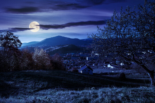 carpathian rural landscape in autumn at night. village in the valley at the foot of the mountain. beautiful countryside scenery in full moon light. trees in fall foliage on the grassy hills