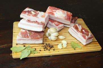 On a wooden board are pieces of lard with a layer of meat, a head of garlic, pepper and bay leaf. Salo recipe. Ukrainian, Russian, German cuisine