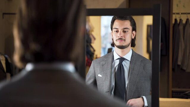 Mirror reflection of young bearded man trying on suit in fashion store.