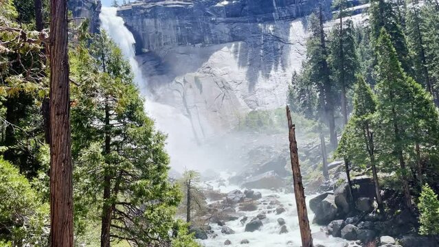 A man takes a picture of the misty waterfall in Yosemite National Park. Vibrant trees and smooth rocks surround the powerful waterfall on a warm spring day.