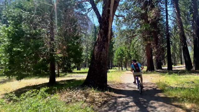 Wider angle of two boys biking on a trail in Yosemite National Park. They pass numerous trees with mountains in the distance. They are on a rocky trail.