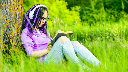 Young woman in glasses, headphones reading book, sitting near tree in summertime. Relaxed secluded...