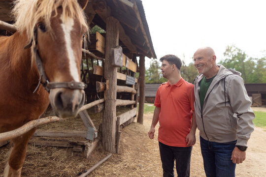 Happy Senior Father With His Adult Son With Down Syndrome At Ranch Looking At Horse.