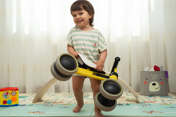 Female toddler holding her yellow tricycle while is laughing and running to the camera. Playful and...